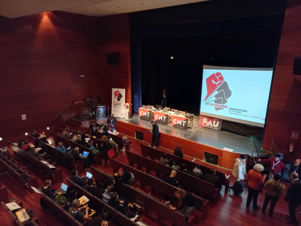 Imagen que muestra el plenario del Congreso Confederal, en el que se ven banderas de la CNT, la FAU, y el logo del congreso proyectado.